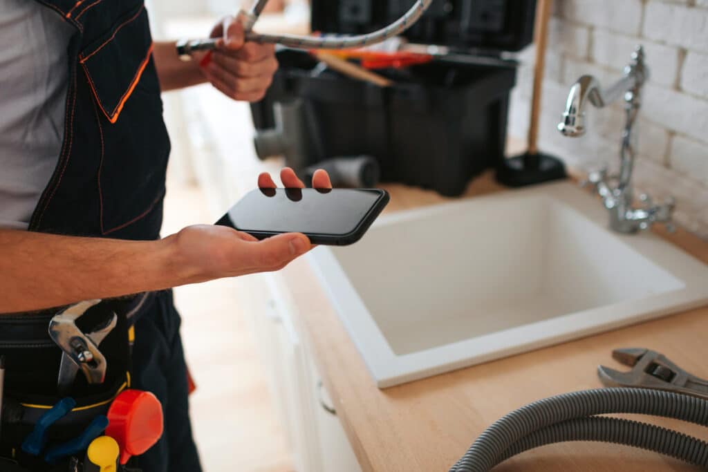 man standing in kitchen at the sink, holding a phone and wrench
